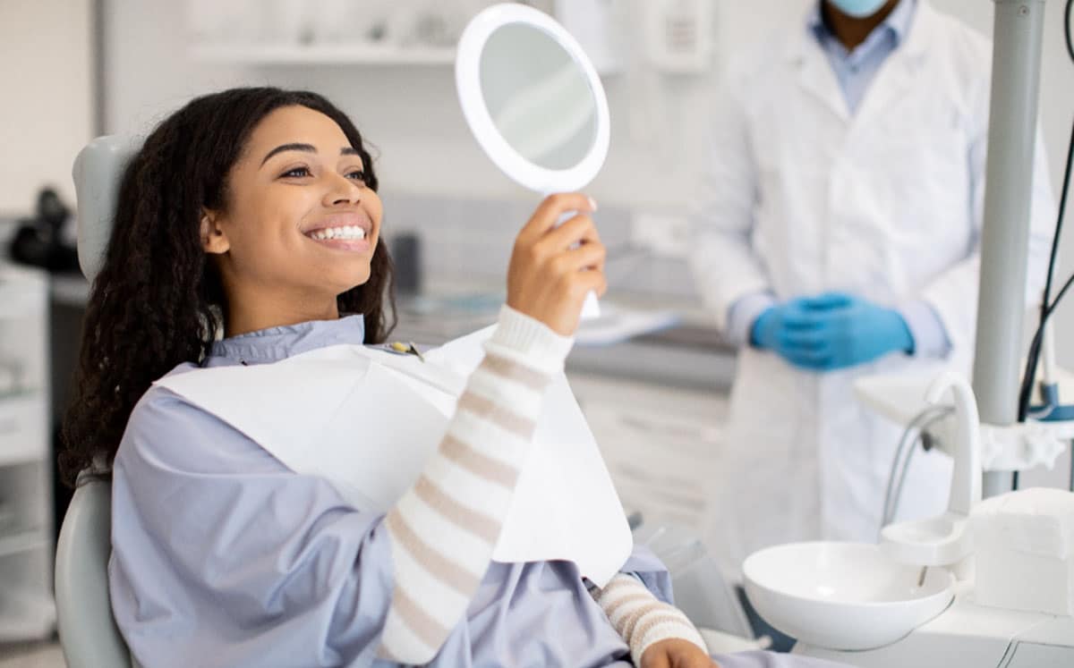 Female dental patient smiling while holding dental mirror in modern dental office with dentist.