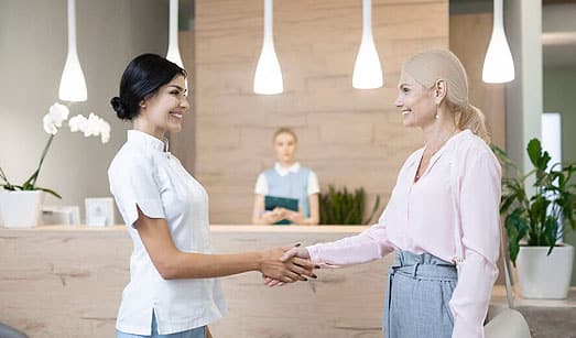 Two women shaking hands in modern dental office reception area with pendant lights and plants.