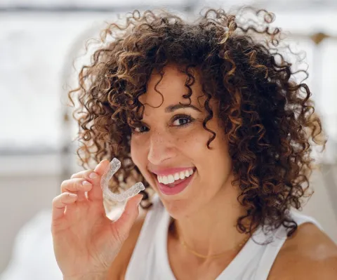 Woman with curly hair smiling while holding dental floss in bright bathroom setting.